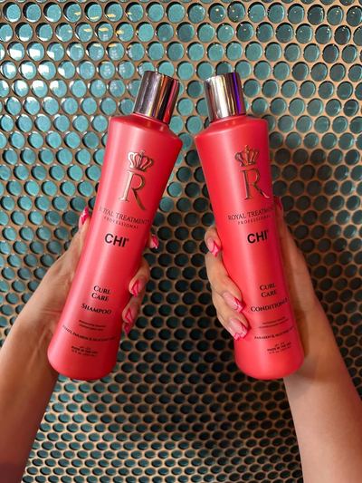 Zwei rote CHI Royal Treatment Curl Care Flaschen werden vor einer türkisfarbenen Fliesenwand in zwei Händen gehalten.