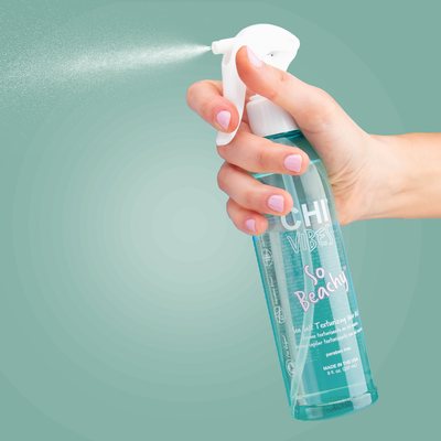 Eine Hand sprüht aus einer türkisfarbenen CHI Vibes So Beachy Flasche vor grünem Hintergrund.
